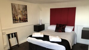 A neatly made bed with white linens and a black headboard, a bedside table with a box of toiletries, and a painting of a city scene on the wall.