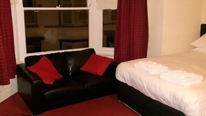 A hotel room with a black leather sofa, a bed with white linens, and red curtains.