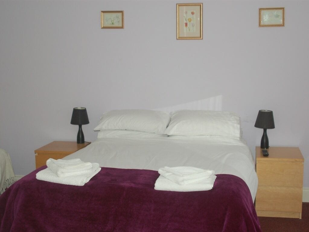 A neatly made bed with white linens, a purple blanket, and two bedside lamps on wooden nightstands.