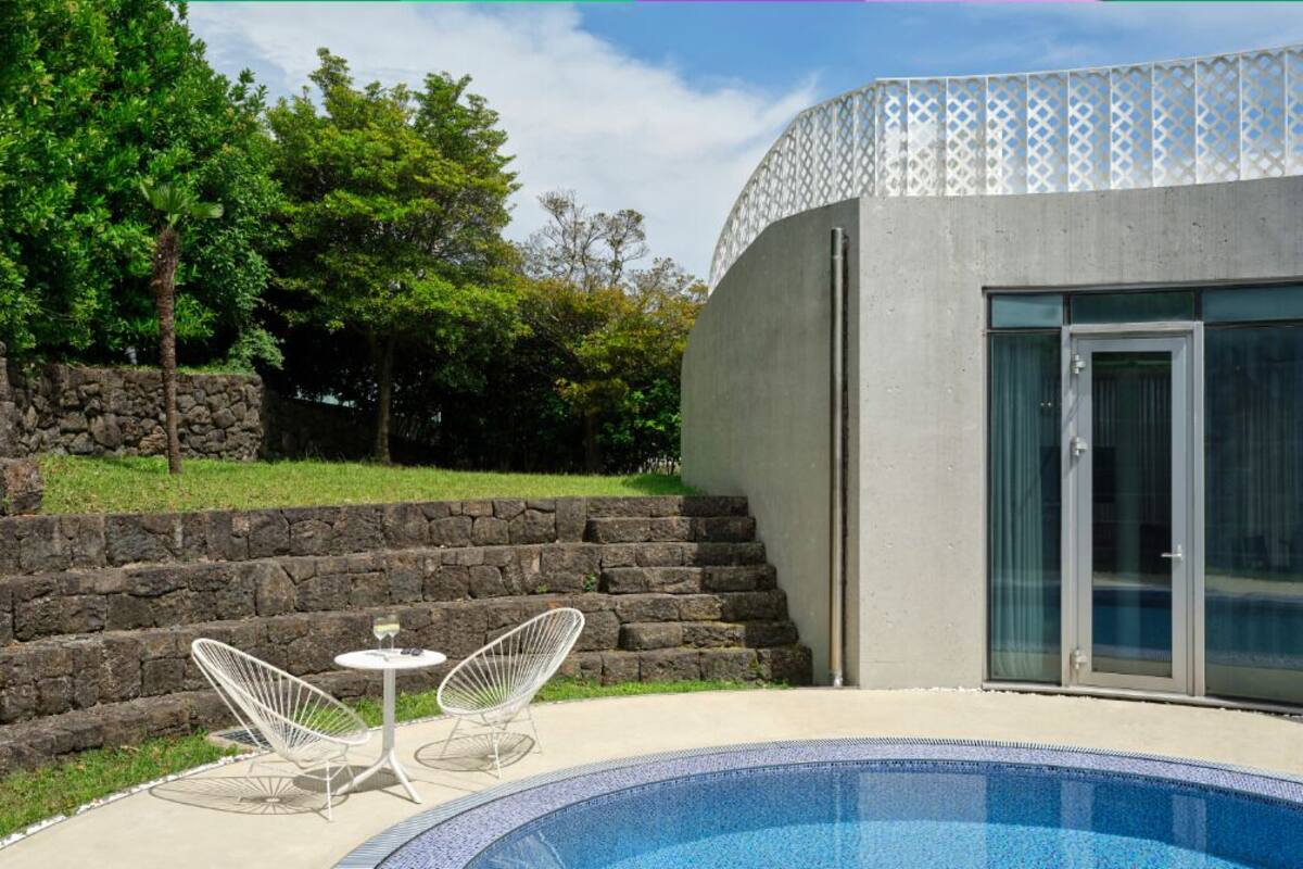 A modern outdoor swimming pool with two white chairs and a small table, surrounded by a stone wall and greenery.