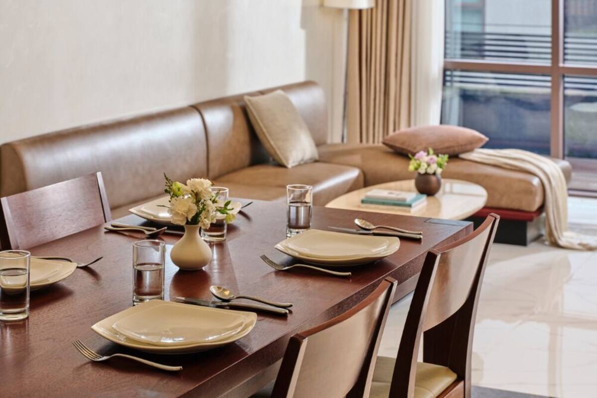 A dining area with a wooden table set for four, surrounded by chairs. There is a sofa and a small round table with a vase of flowers.
