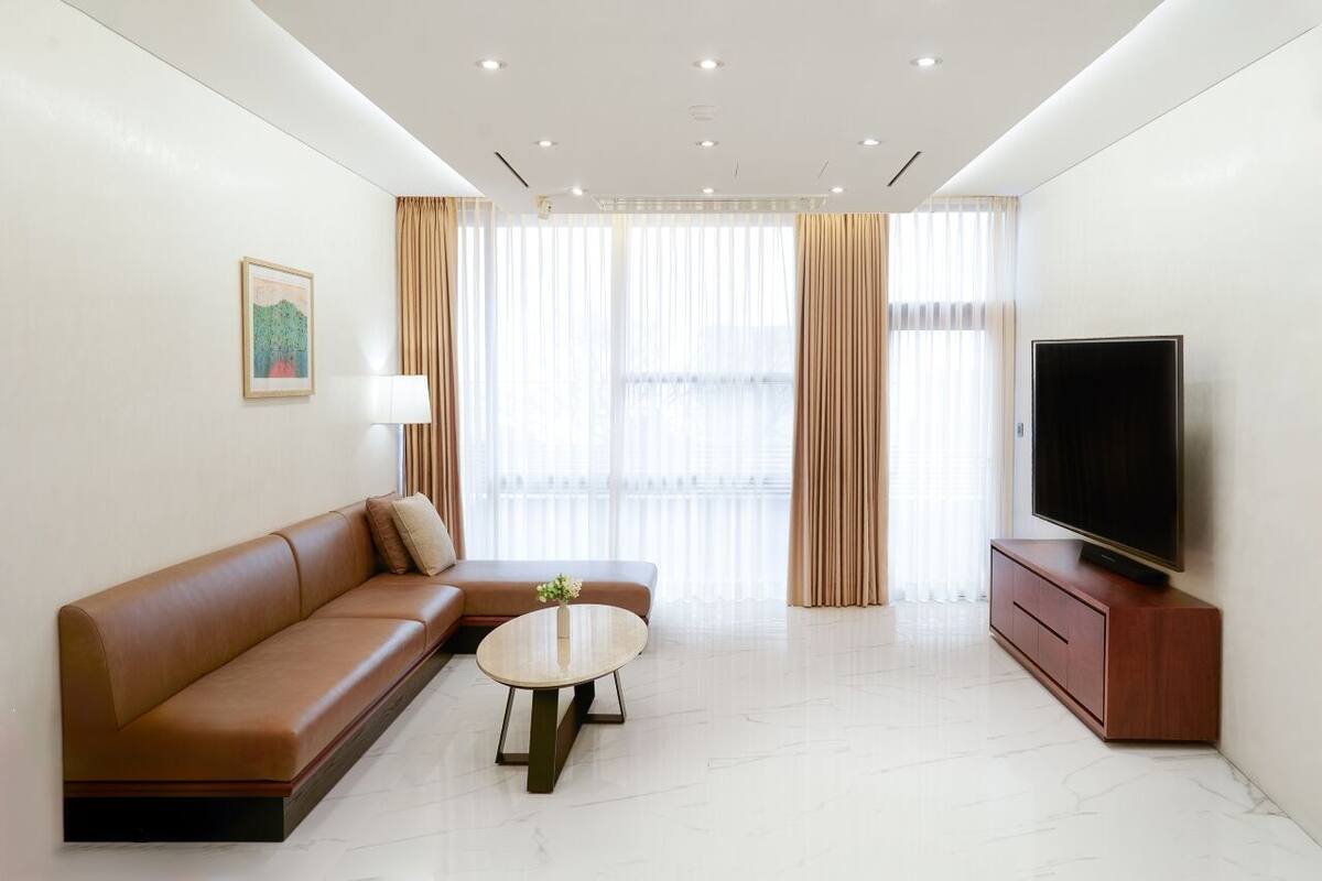 A modern living room with a brown leather sofa, a round coffee table, a flat-screen TV on a wooden cabinet, and large windows with sheer curtains.