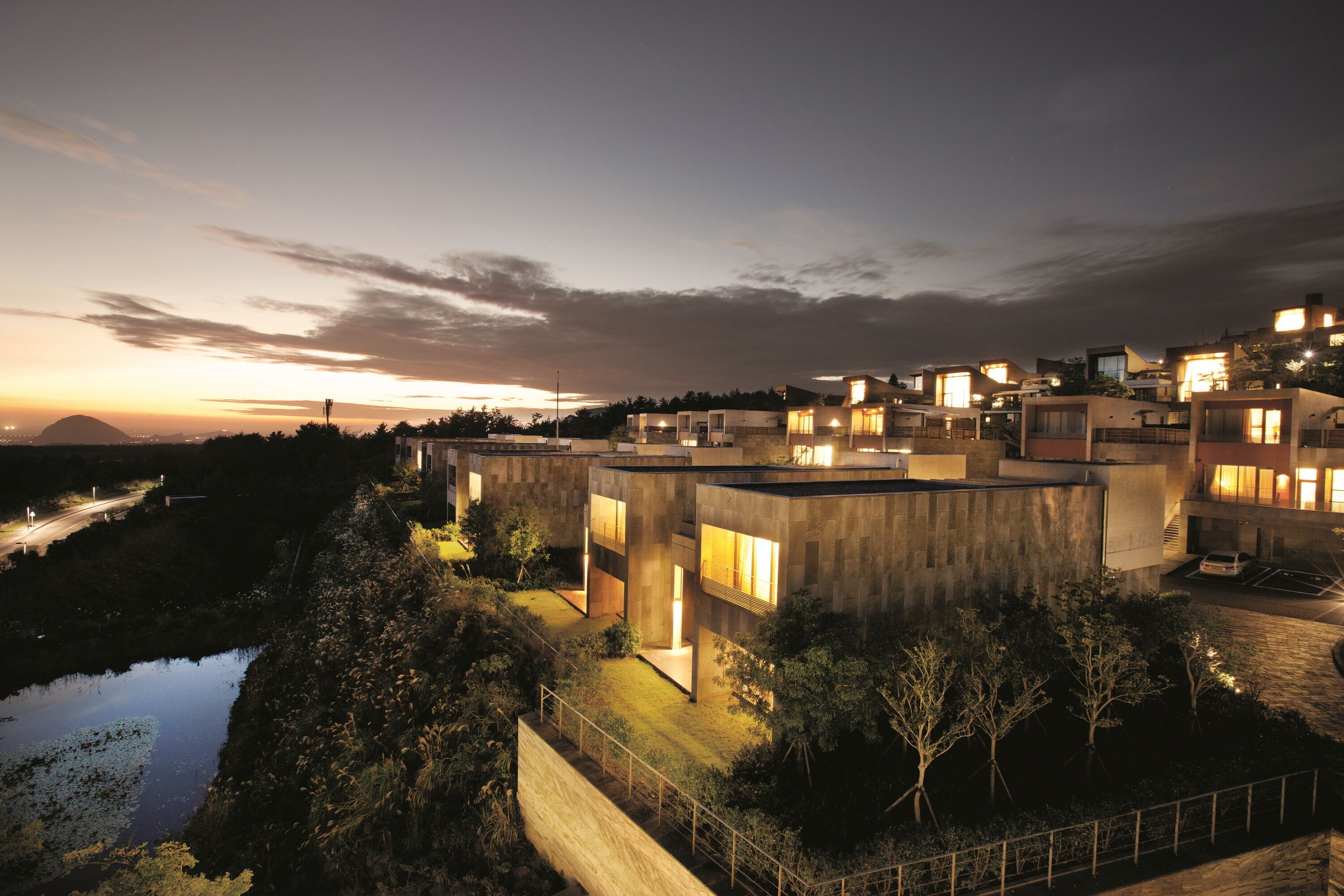 A modern residential complex with multiple buildings, a visible water body, and a road in the background.