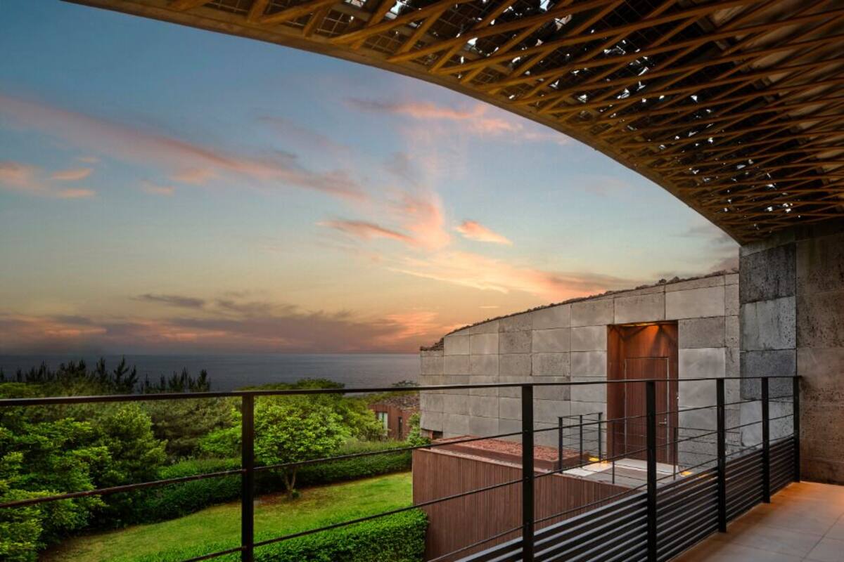 A balcony with a view of the sunset over the ocean, a stone wall, and a wooden pergola.