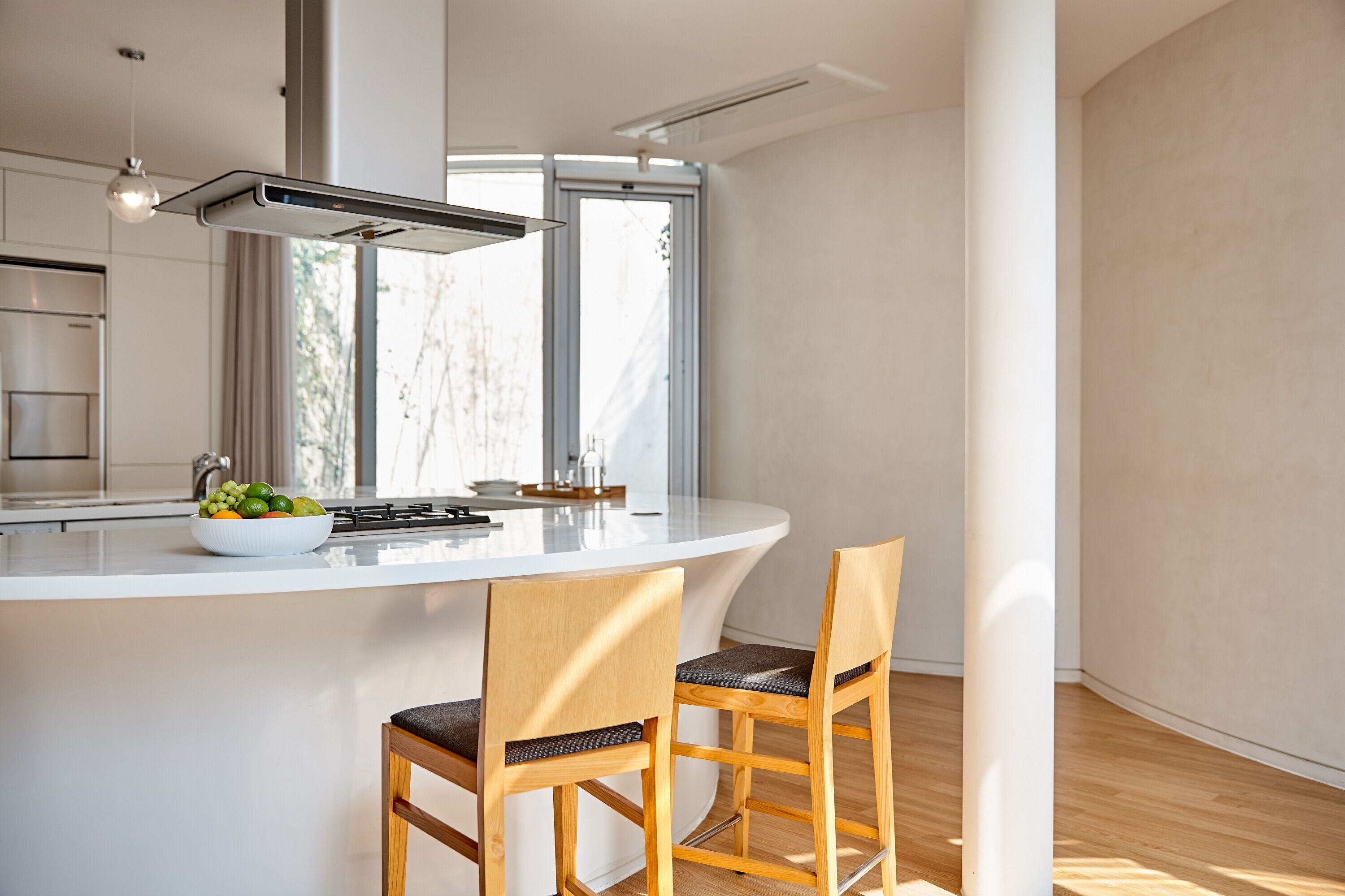 A modern kitchen with a white island, two wooden chairs, a bowl of fruit, and a window providing natural light.