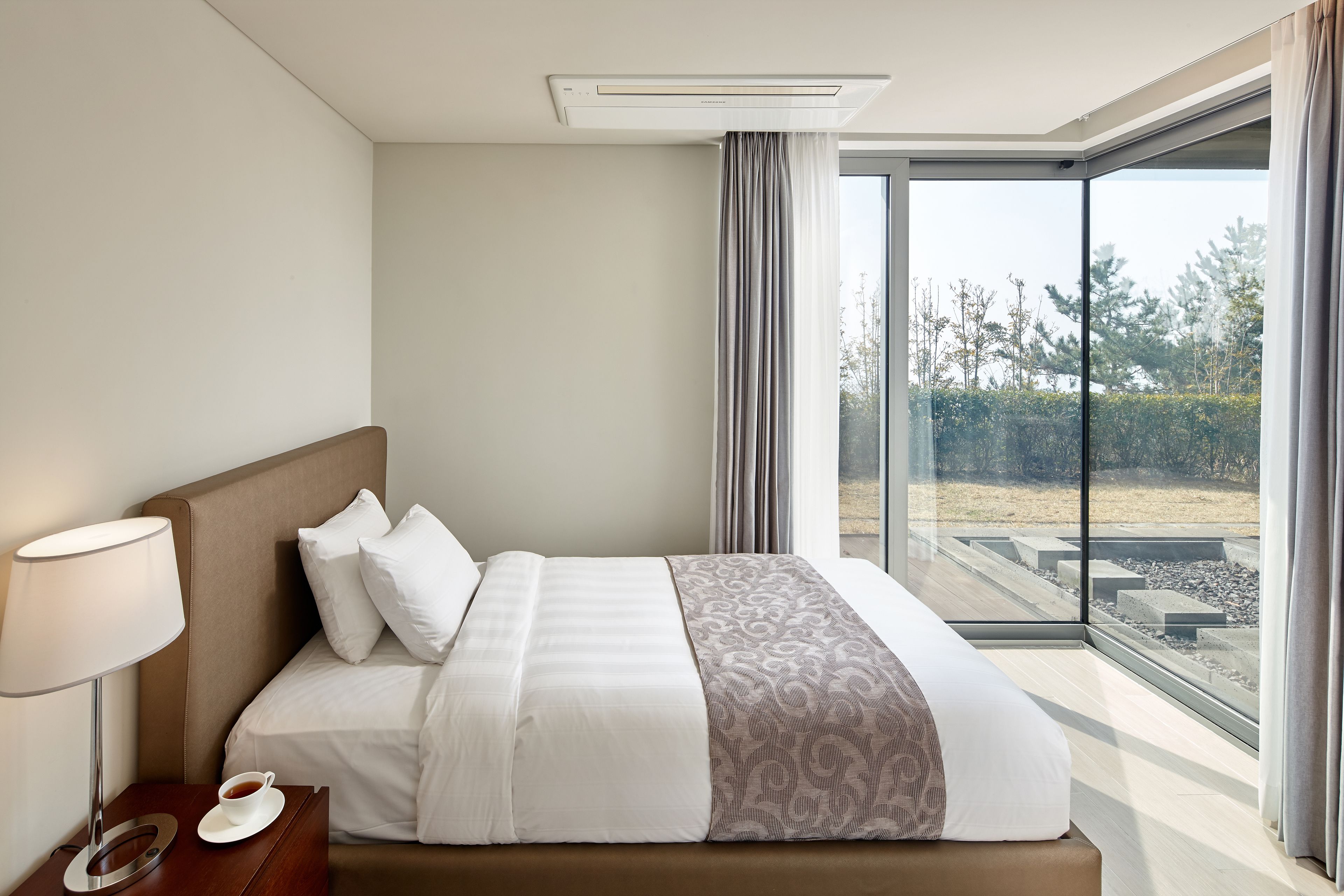 A hotel room with a large bed, a bedside table with a lamp, and a view of the outdoors through a large window.