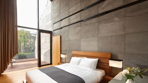 Habitación de hotel moderna con una cama grande, un cabecero de madera, una mesita de noche con lámpara y vistas al exterior a través de una puerta corredera.
