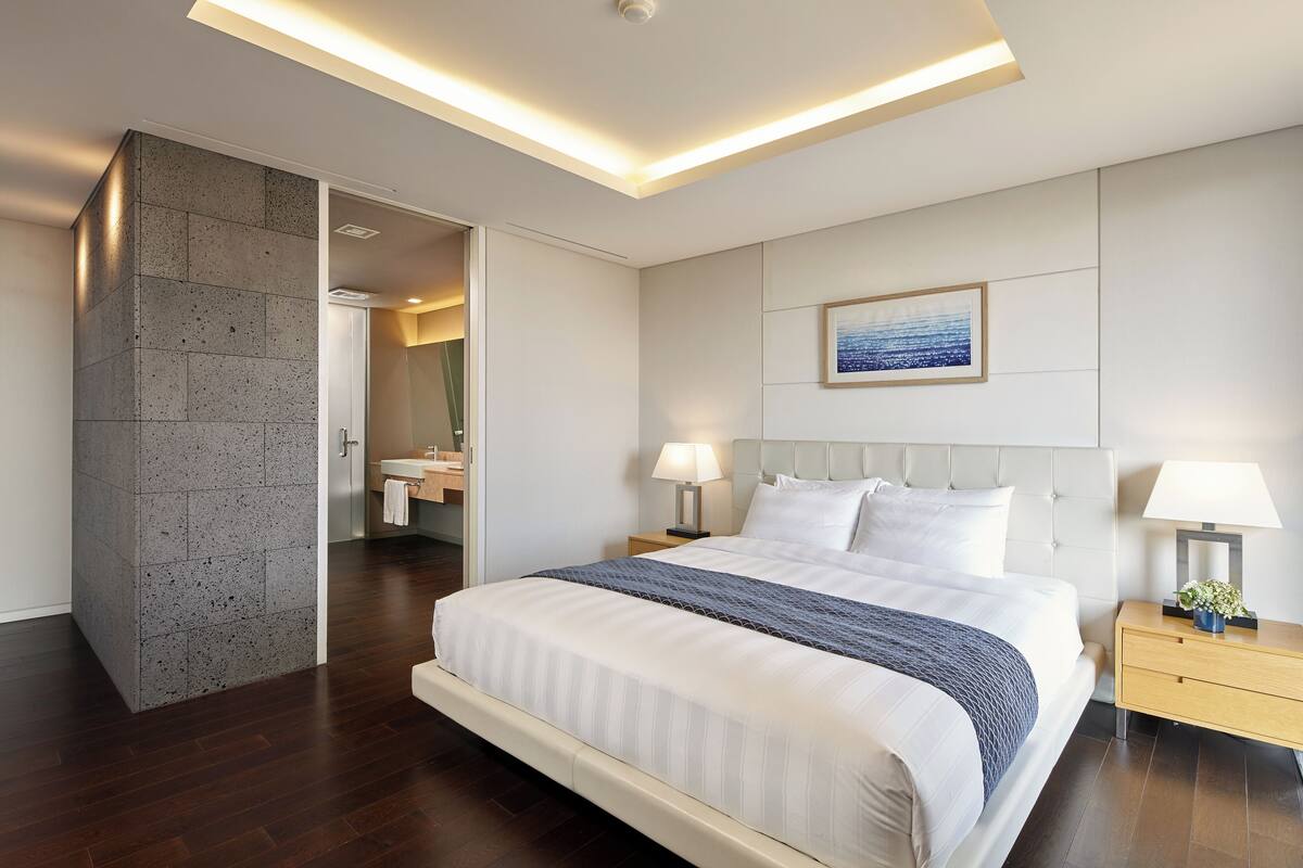 A modern hotel room with a large bed, bedside tables, a nightstand, a bathroom with a mirror, and a painting on the wall.
