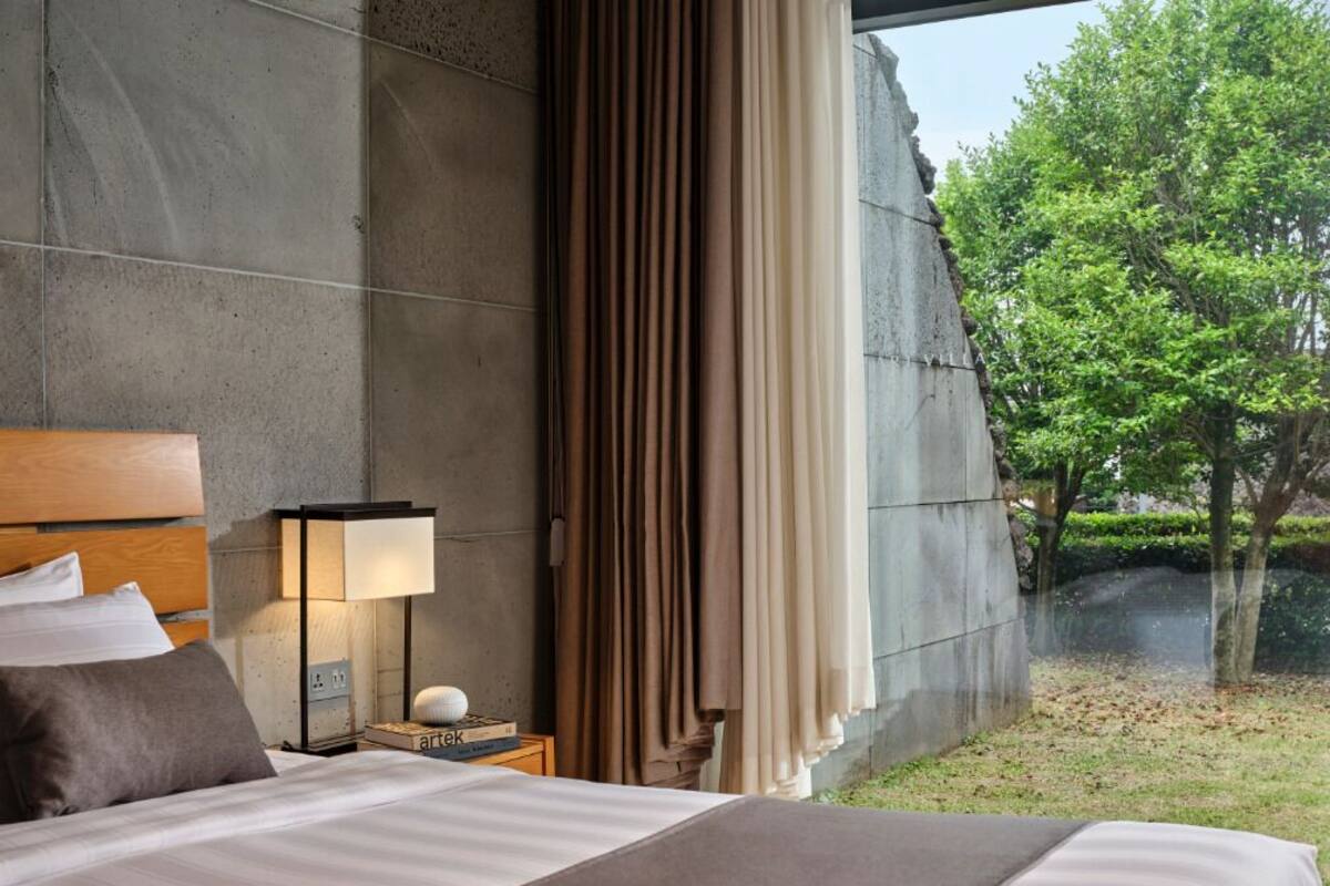 A modern hotel room with a large bed, a nightstand with a lamp, and a view of a garden and trees.