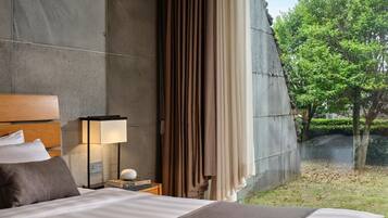 A modern hotel room with a large bed, a nightstand with a lamp, and a view of a garden and trees.