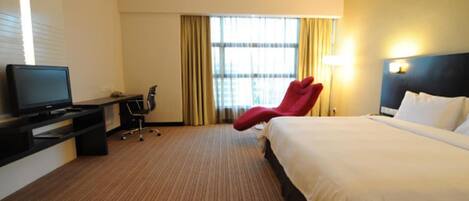 Una habitación de hotel con una cama, un escritorio con televisor, una silla roja y una ventana con cortinas.