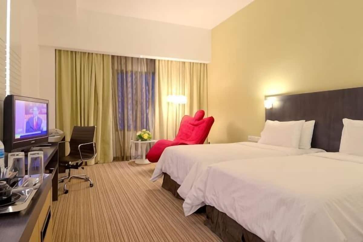 A hotel room with a bed, a desk with a television, a red chair, and a small table with a lamp.