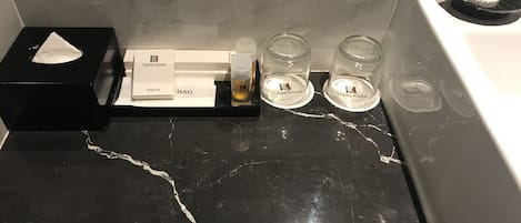 Una encimera de baño con superficie de mármol negro, una bandeja negra que contiene una caja de pañuelos, un gorro de ducha y una bolsa con la etiqueta "THEKLAGAN". También hay dos botellas de vidrio transparente con el mismo nombre de marca.