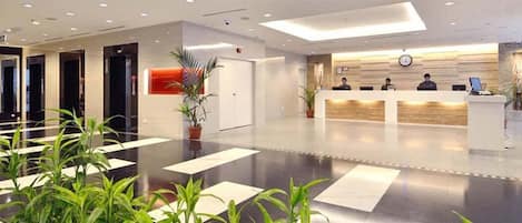 A modern hotel lobby with a reception desk, several receptionists, and a large potted plant.