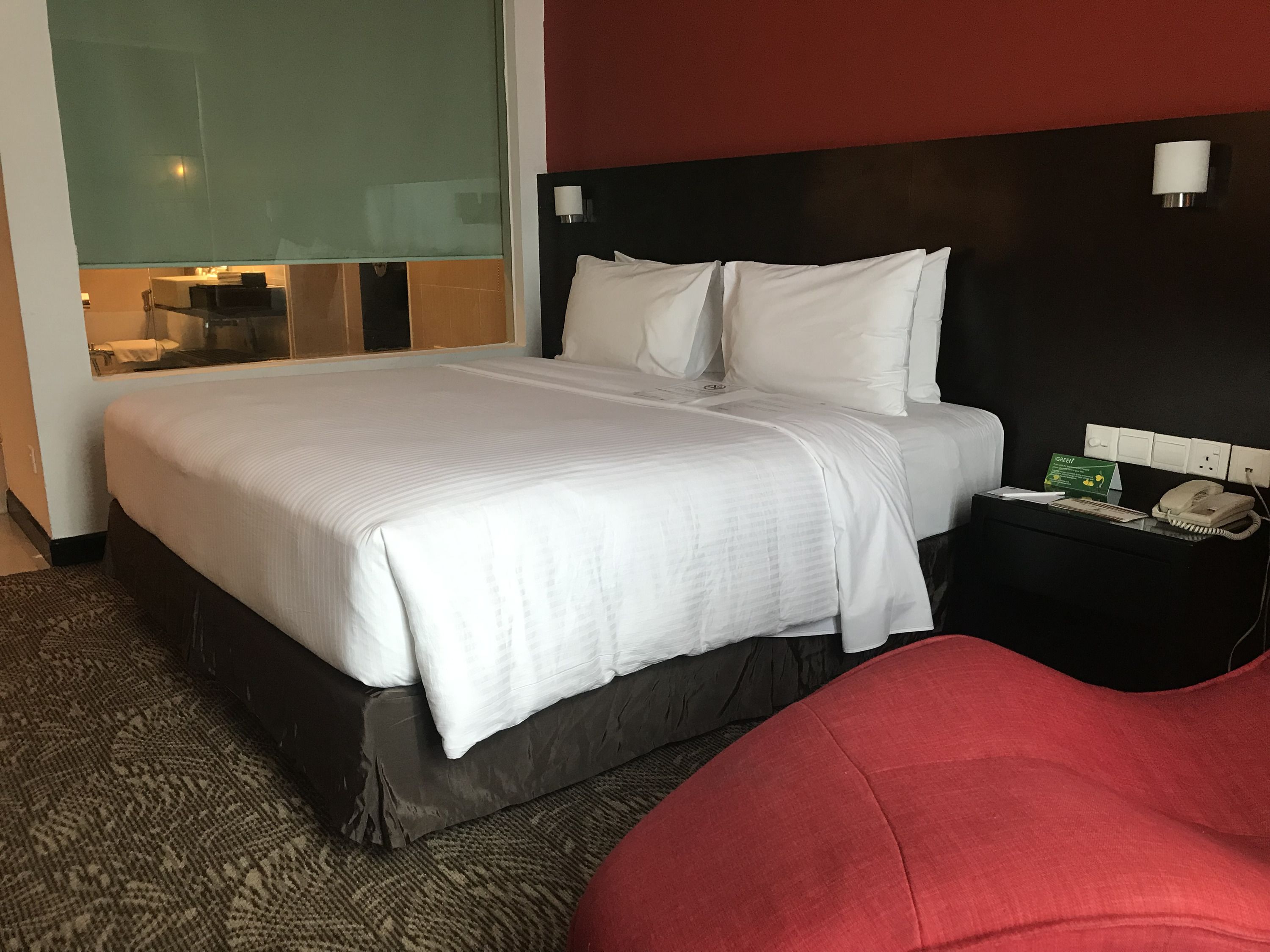 A hotel room with a large bed, a bedside table with a telephone, and a glass partition separating the room from the hallway.
