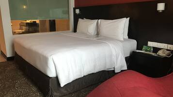 A hotel room with a large bed, a bedside table with a telephone, and a glass partition separating the room from the hallway.