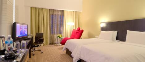 Una habitación de hotel con una cama, un escritorio con televisor, una silla roja y una mesita de noche con lámpara.