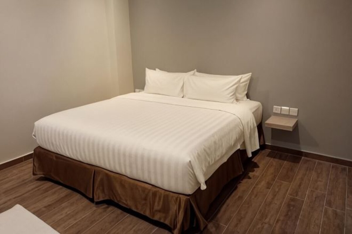 A neatly made bed with a brown bed skirt, a bedside table with a lamp, and a wall-mounted light switch.