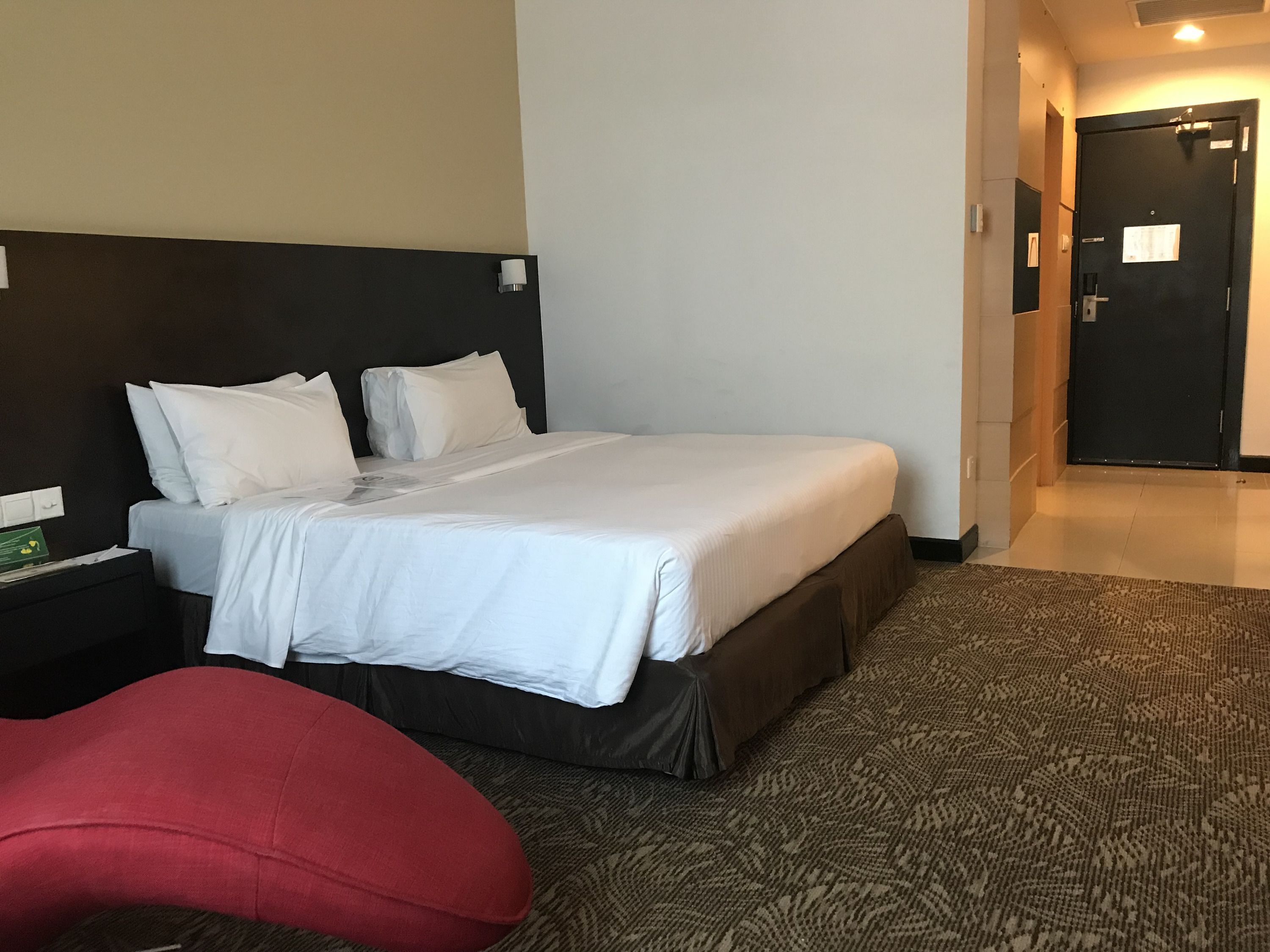 Habitació d'hotel amb un llit gran, una tauleta de nit, una cadira vermella i una porta que dona a una altra habitació.