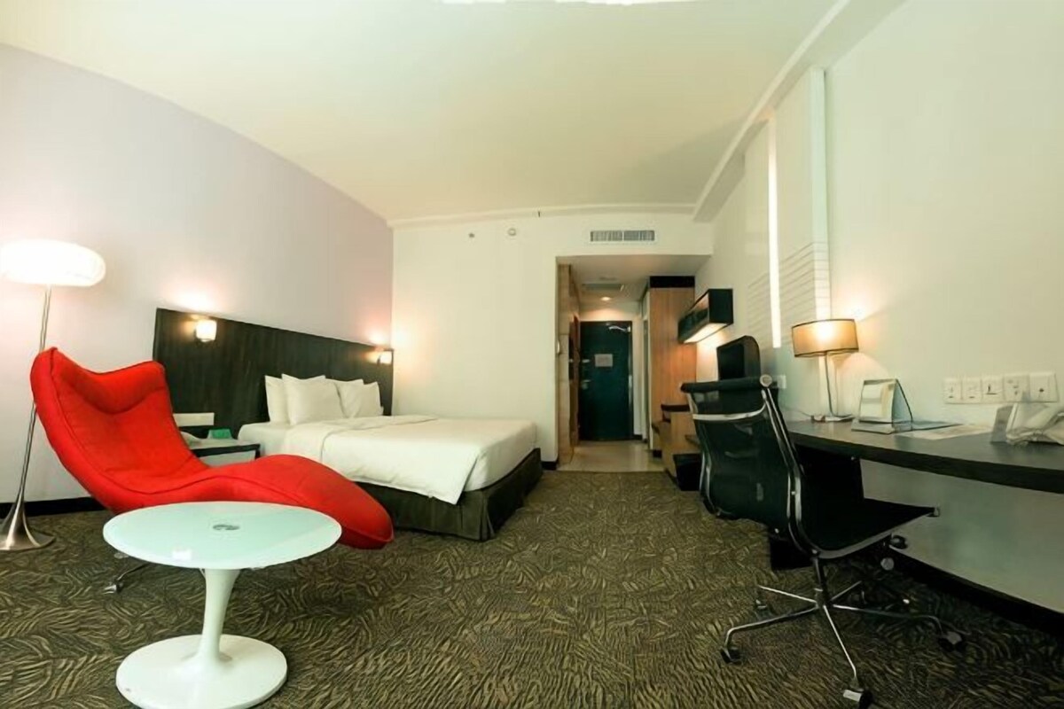 A hotel room with a bed, a red chair, a small table, a desk, and a lamp.