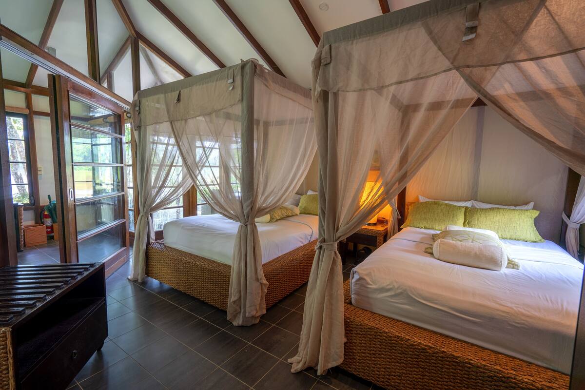 A room with two beds, each with a canopy and a nightstand. The room has a large window and a view of greenery outside.