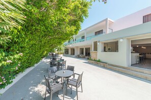 Un patio con mesas al aire libre, un porche cubierto y un edificio con varios balcones y una zona de cocina.