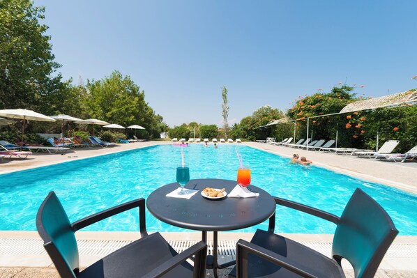 A poolside view with lounge chairs, umbrellas, and a table with drinks and a plate of food.