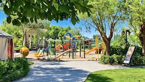 Un parque infantil con diversas estructuras de juego, un espacio de descanso techado y un camino pavimentado.