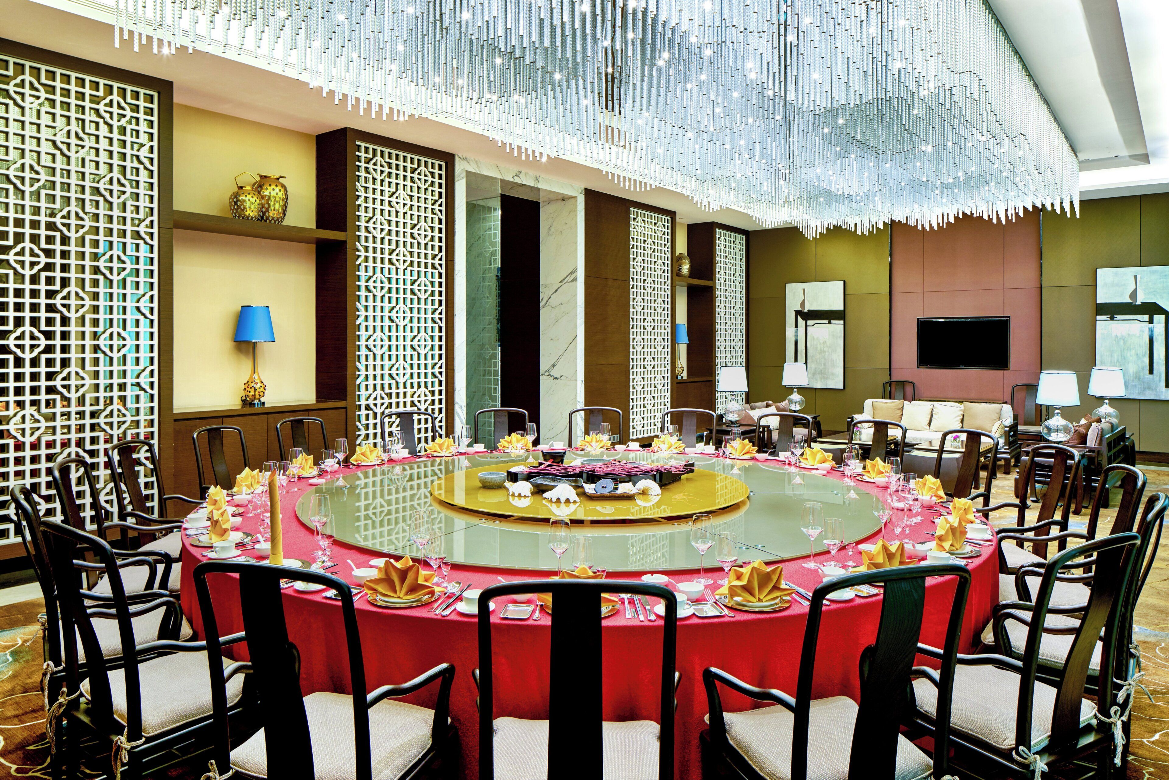 Una mesa redonda lista para una comida, con sillas alrededor, un techo decorativo y un televisor en la pared.