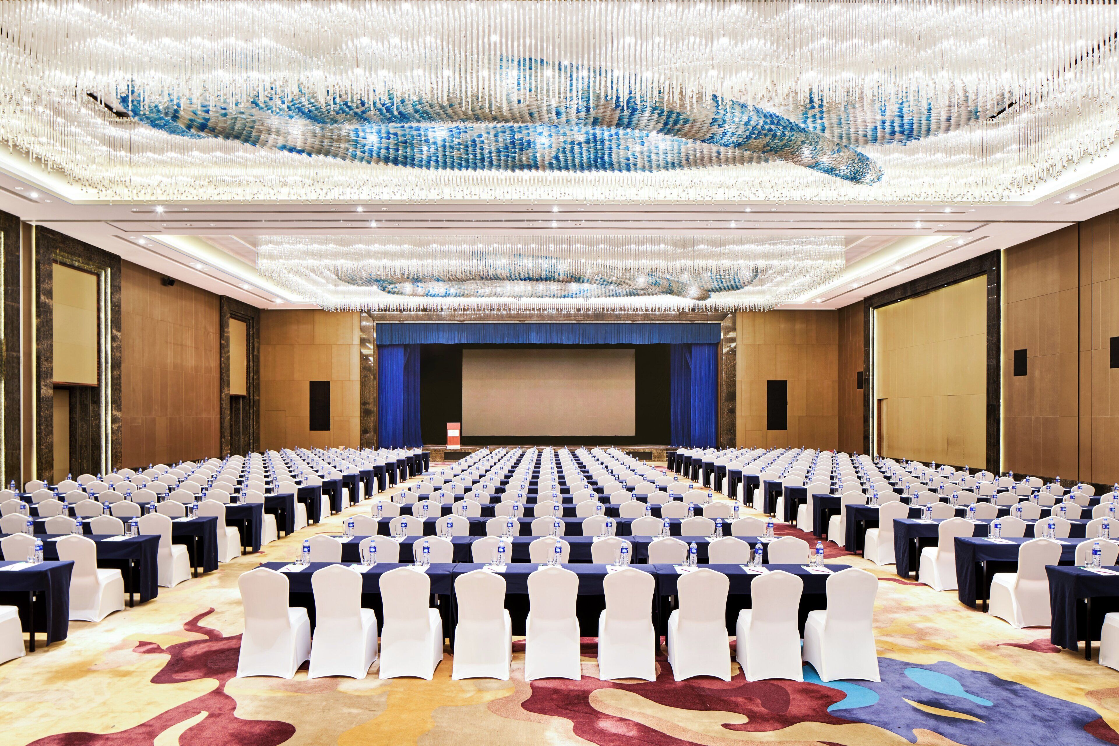 Un salón de conferencias con filas de mesas y sillas, una pantalla grande al frente y un candelabro colgando del techo.