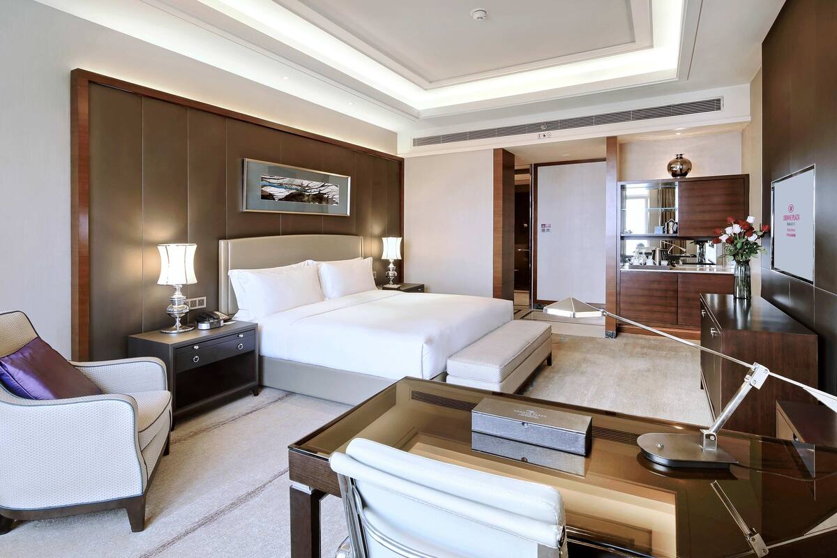 A hotel room with a large bed, a desk with a lamp, a chair, a bedside table, and a mirror.