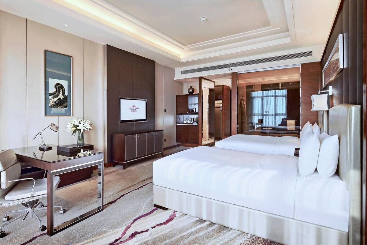 A modern hotel room with a large bed, a desk with a chair, a television, and a bathroom visible through a glass partition.