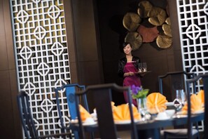 A restaurant interior with a waiter holding a tray, tables with orange napkins, and a decorative wall with circular patterns. - Crowne Plaza Tianjin Jinnan by IHG (Tianjin)