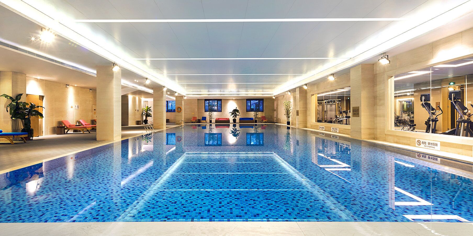 Una amplia piscina cubierta con suelo de azulejos azules, rodeada de sillones de descanso y equipo de ejercicio.