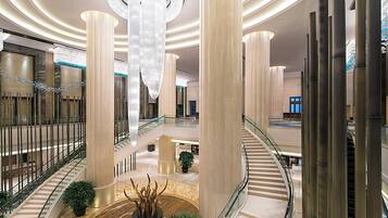 A spacious lobby with a grand staircase, high ceilings, and a central chandelier.