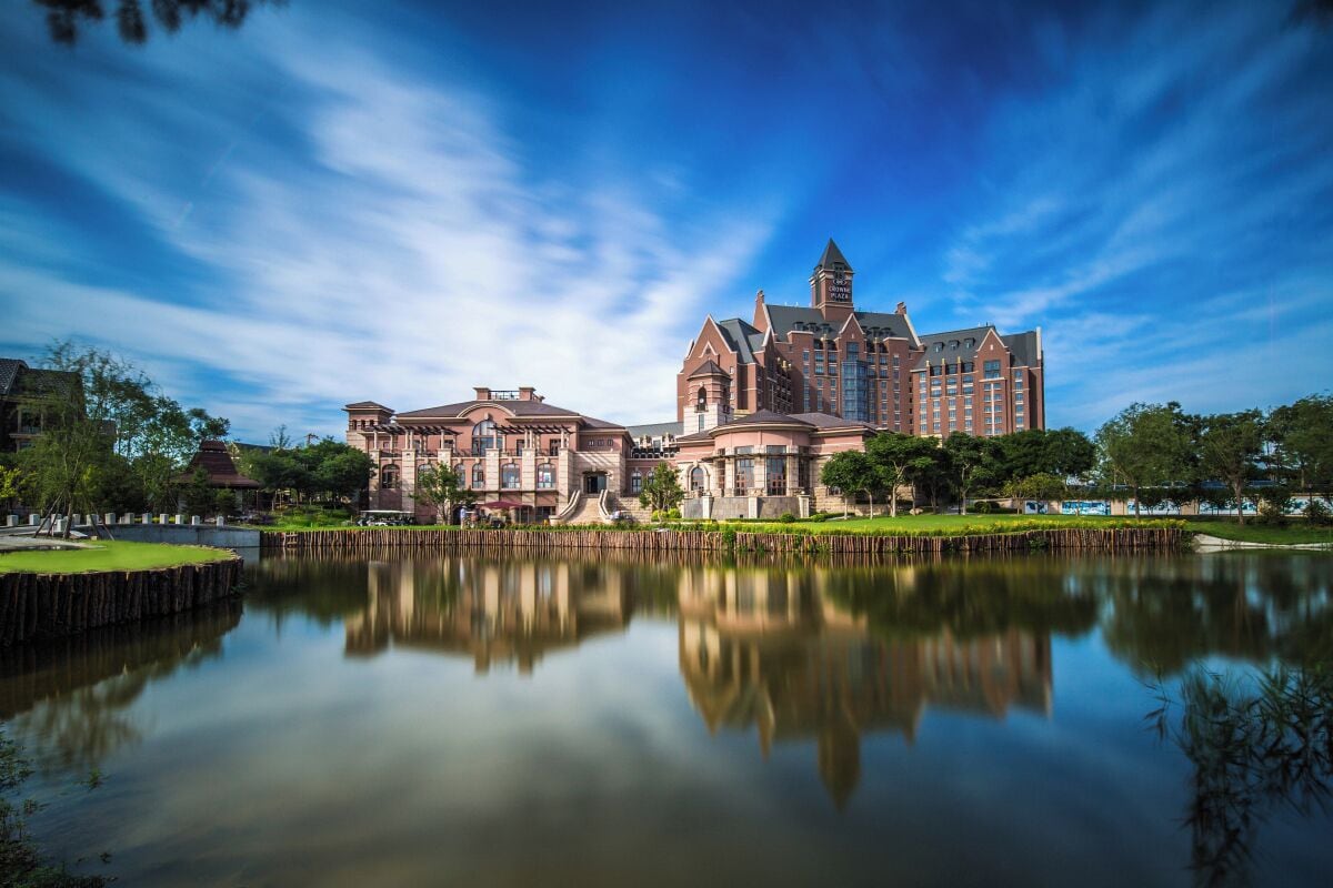 A large hotel with a reflection in a calm body of water, surrounded by greenery.