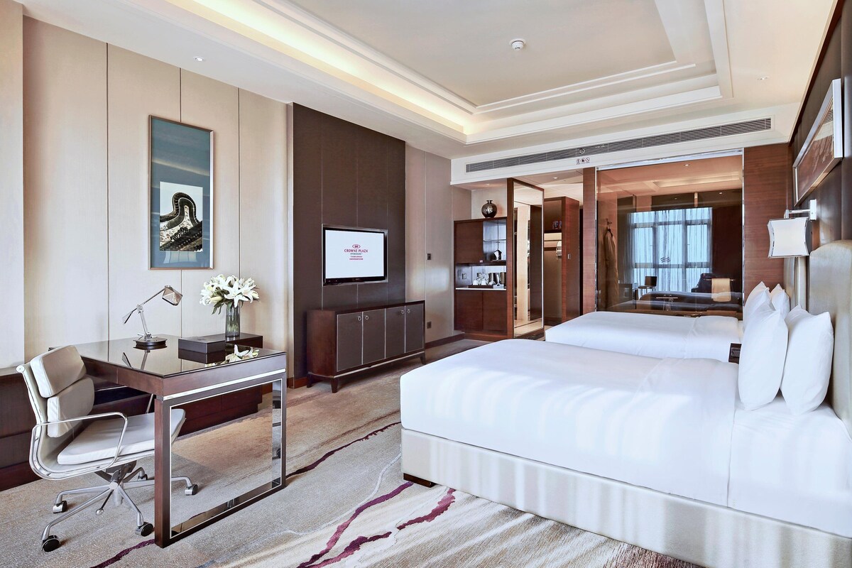 Habitación de hotel moderna con una cama grande, un escritorio con silla, televisión y baño visible a través de un panel de vidrio.