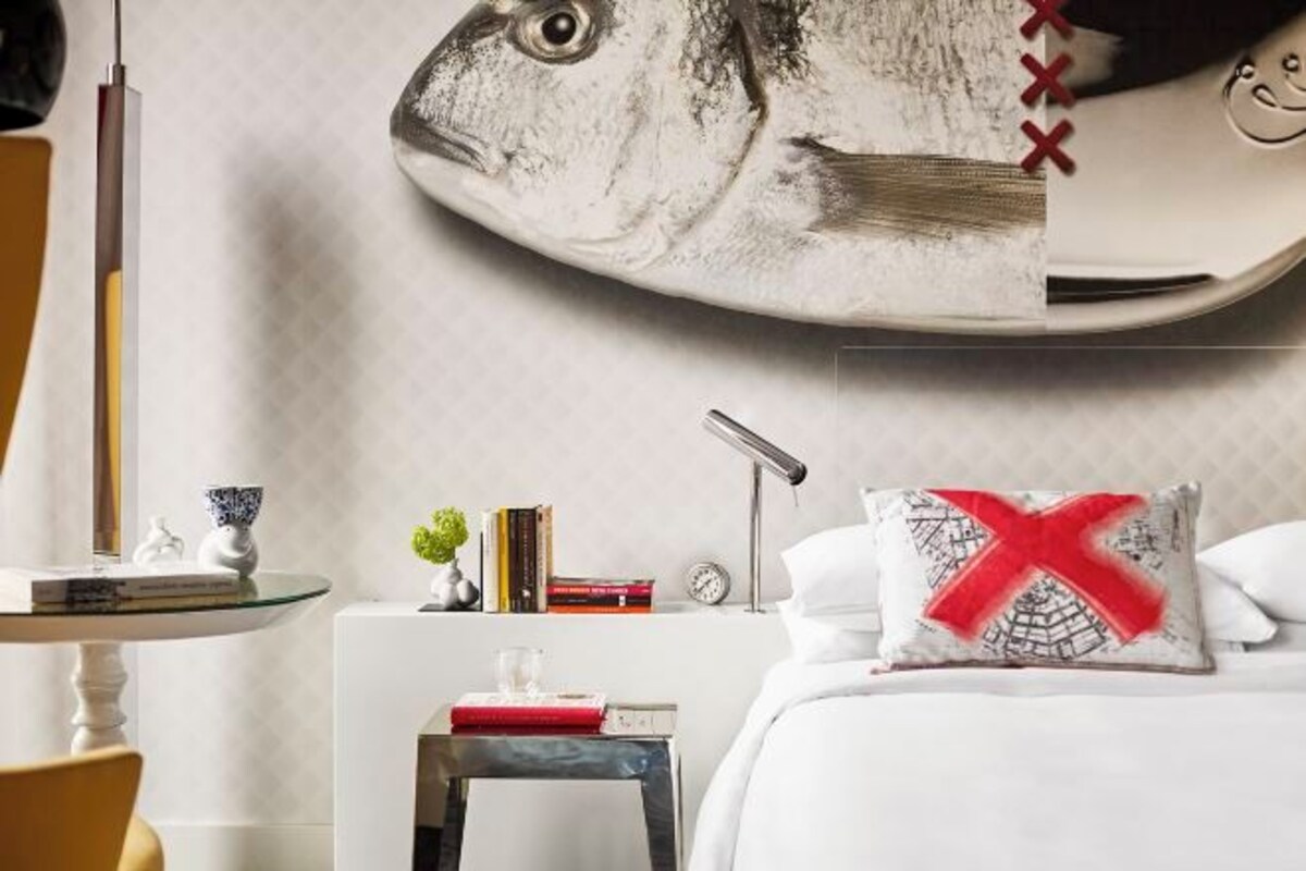 A bedroom with a large fish mural on the wall, a bedside table with books, a lamp, and a pillow with a red 'X'.