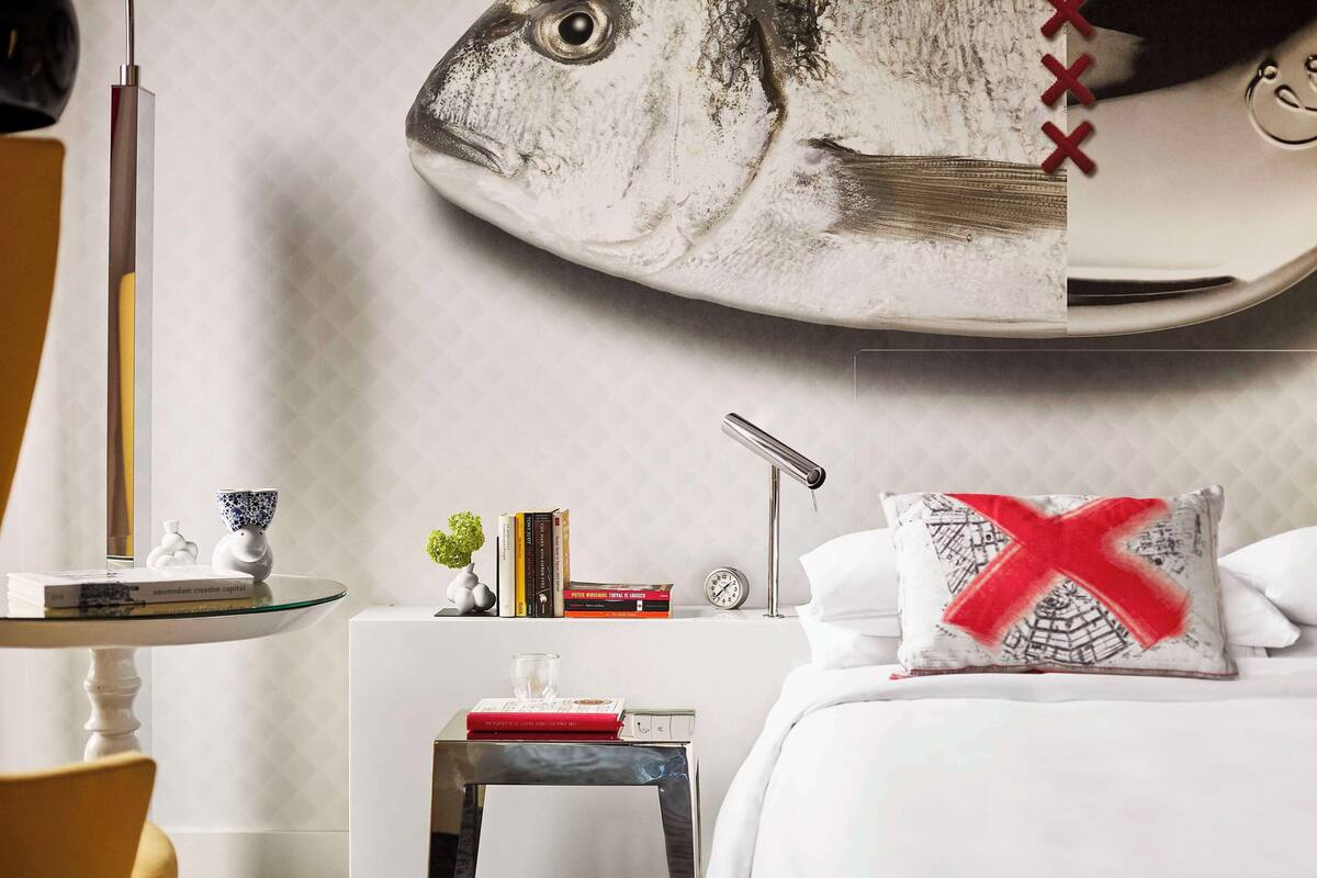 Un dormitorio con cama, mesita de noche, una mesa con libros, lámpara y un mural de peces en la pared.