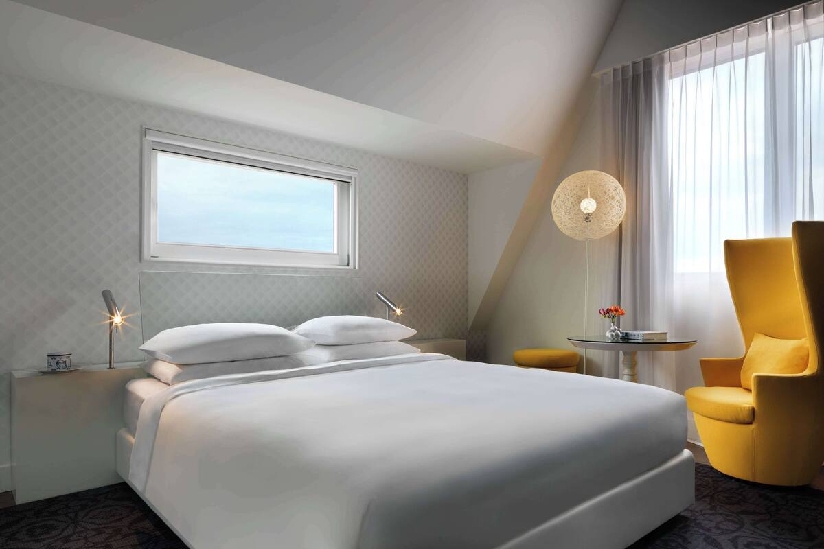 Habitación de hotel con una cama grande, una mesita de noche con lámpara, una silla y una pequeña mesa con un jarrón.