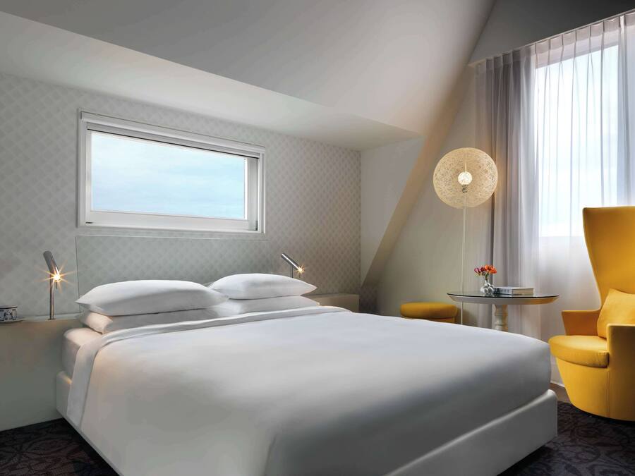 Una camera d'albergo con un letto grande, un comodino con una lampada, una sedia e un tavolino con un vaso.