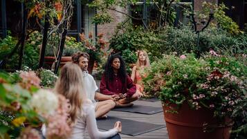 A group of people sitting on yoga mats in a garden with potted plants and a brick building in the background.