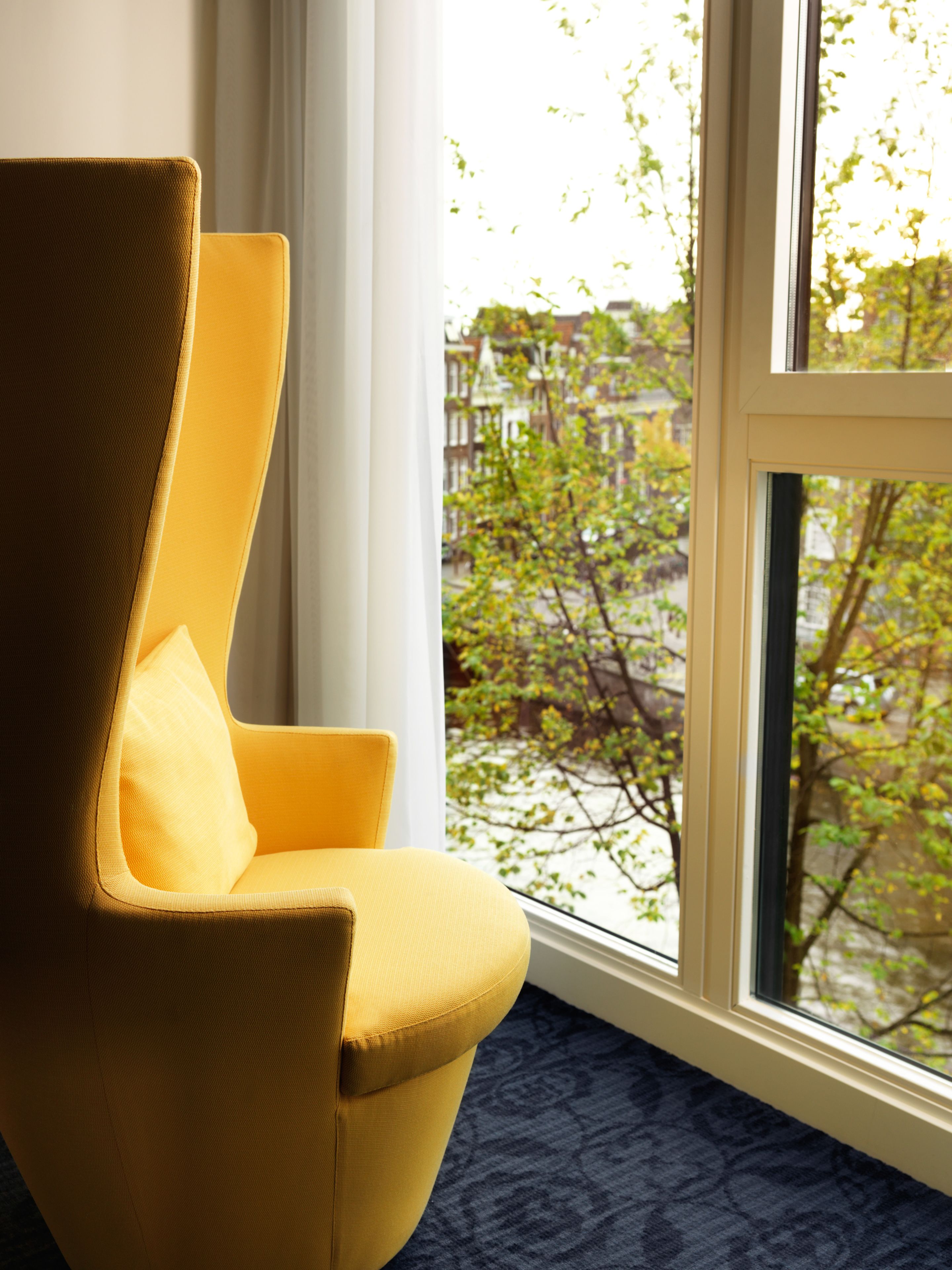 Un sillón amarillo junto a una ventana con vistas a un árbol y edificios.