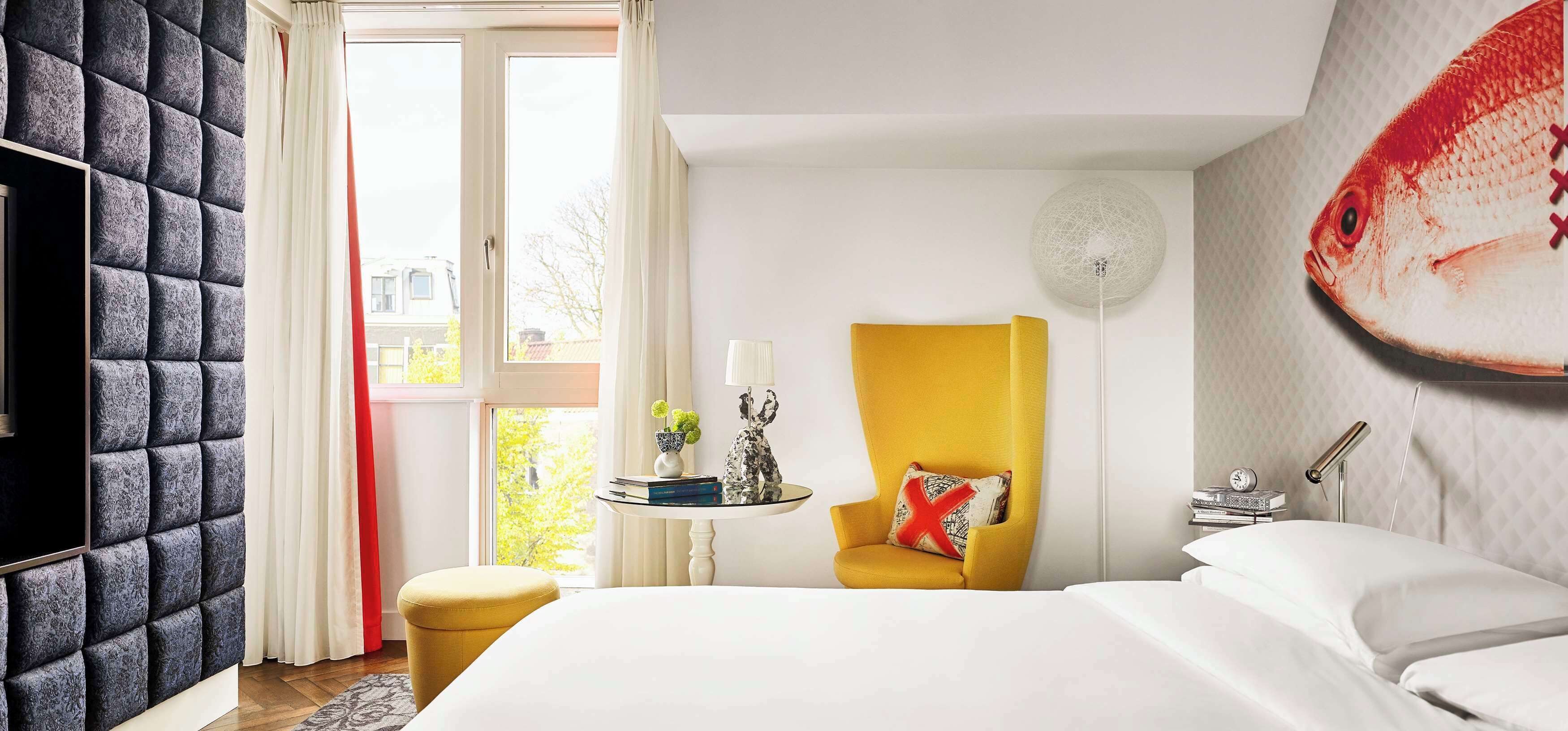 Habitación de hotel con cama, un sillón amarillo, una mesa redonda con lámpara y un mural de peces en la pared.