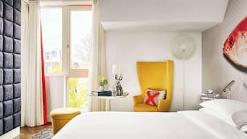 Habitación de hotel con cama, un sillón amarillo, una mesa redonda con lámpara y un mural de peces en la pared.