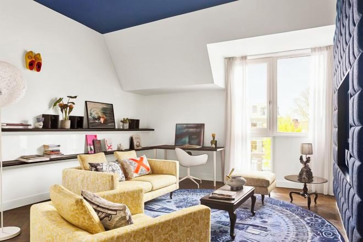 A living room with a sofa, coffee table, and a desk with a chair. There is a large window with curtains, a lamp, and a wall-mounted TV.