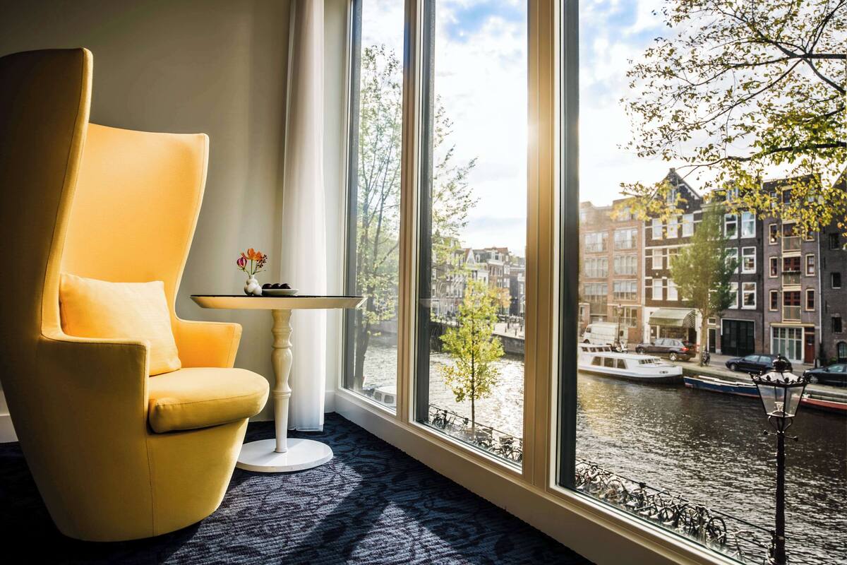 Una habitación con un ventanal grande, un sillón amarillo, una mesita con un florero, y vistas a un canal con edificios.