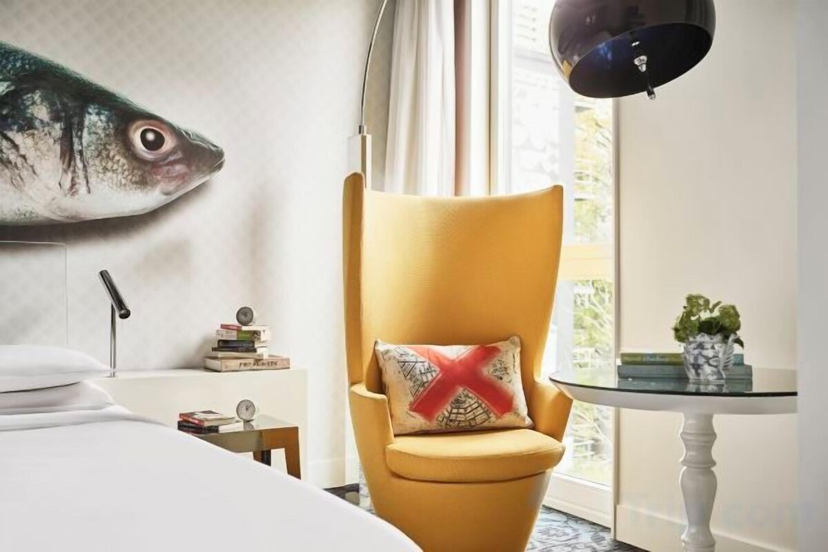A room with a yellow armchair, a white bed, a fish mural, a lamp, and a small table with a plant.
