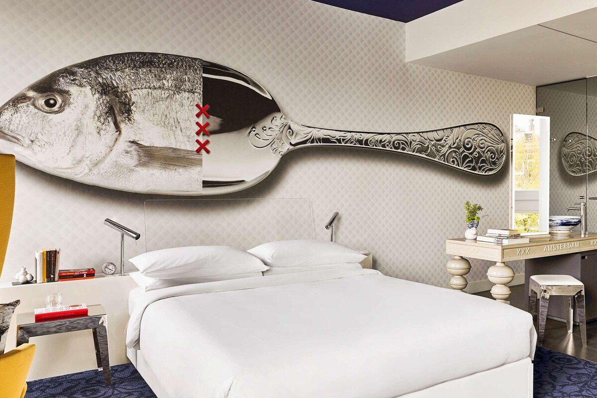A modern hotel room with a large fish mural on the wall, a bed, a desk with a lamp, and a view of the city through the window.