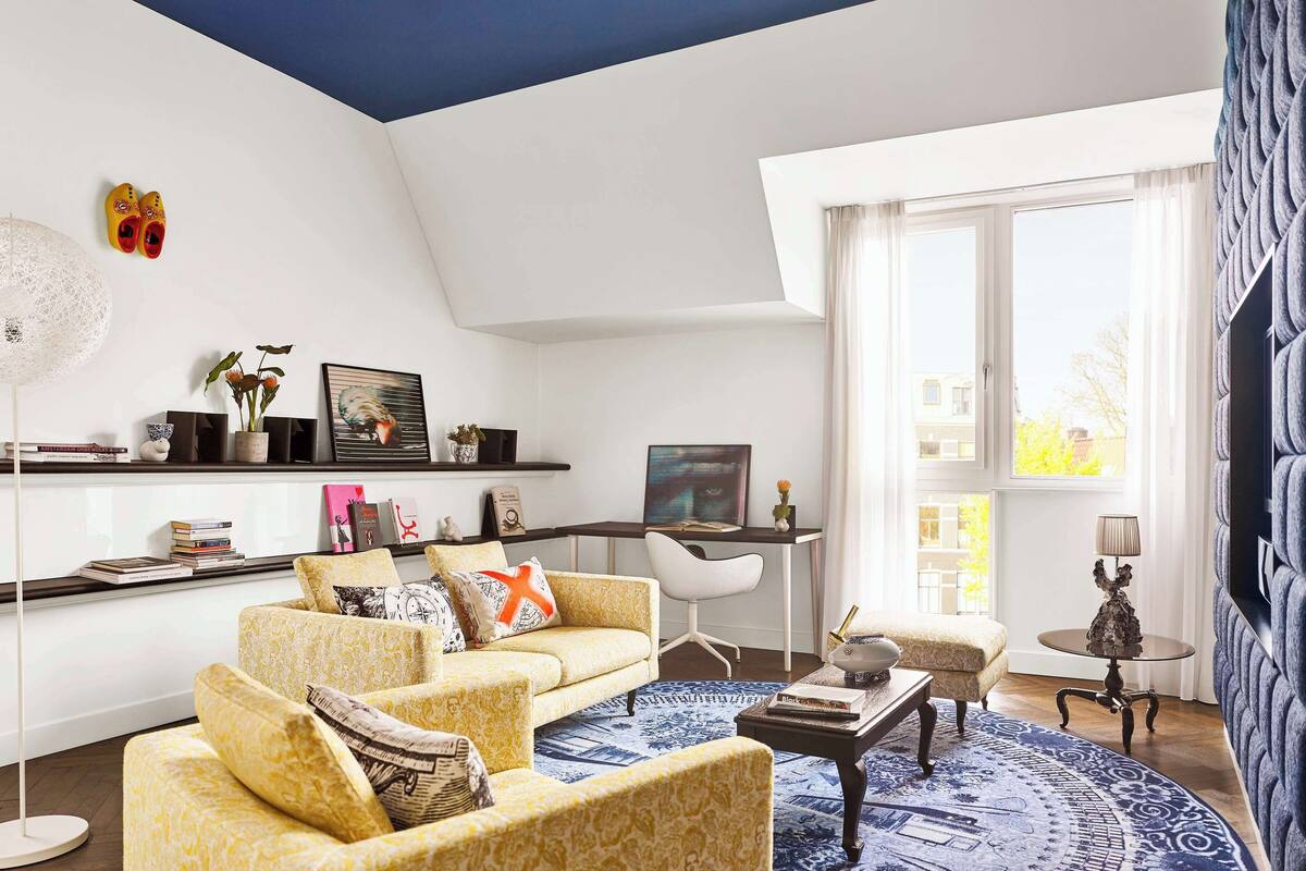Una sala de estar moderna con sofá, mesa de centro y un escritorio con silla. Hay un ventanal con cortinas, una lámpara y un jarrón decorativo sobre una mesita auxiliar.